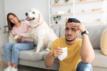 Canvas Print - Unhappy Arab guy suffering from virus or allergy, holding paper tissue, his girlfriend and dog on background, free space