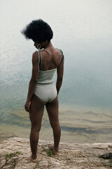 African beautiful model with perfect body in cerulean bodysuit. Voluminous hair, chocolate skin. Healthy looking. Water on background, natural colors. Summer landscape. Calm and balance