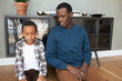 Horizontal portrait of young adult African American man sitting next to his upset son on floor leaning against stylish small bookshelf looking at him, scolding, talking about misbehavior
