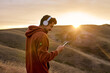 fit caucasian happy man in headphones chatting with someone on smartphone, typing message and smiling outdoors, having rest, enjoy time at sunrise or sunset. in mountains in nature