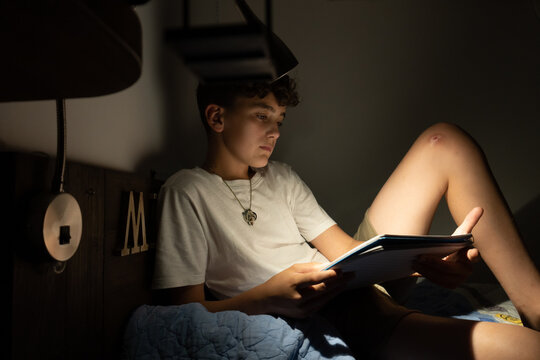 Side view boy in his room lying on his bed reading some notes at night with the illumination of his bedside lamp