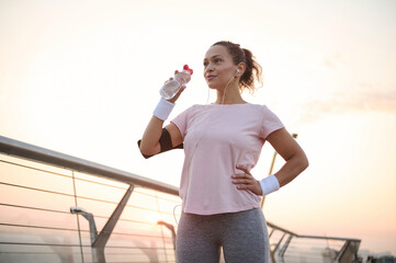 Beautiful sportswoman, female athlete drinks water to hydrate body after outdoor cardio workout. Middle aged athletic woman resting after jogging at dawn. Sport, body care and active lifestyle concept