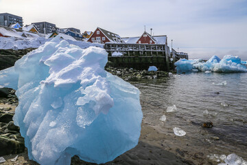 Wall Mural - Big piece of blue ice laying among the stones on the shore with modern Inuit building on the hill at the fjord, Nuuk city, Greenland