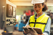 Standing In Front Of A Control Panel, A Female Industrial Electrical Engineer With A Safety Hardhat On Her Head And A Tablet In Her Hand Checks And Maintains CNC Machines In A Factory.