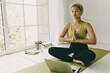 © shurkin_son - Dark-skinned blonde curly-haired female sitting at yoga mat in prayer position and eyes closed, meditating, following online instructor, sitting in front of opened laptop near panoramic window