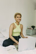 © shurkin_son - Young African stylish girl learning foreign language in bed on lazy morning at home, sitting in front of laptop in earphones, smiling at camera with warm smile, copybook and pen lying on bed