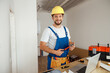 © Kostiantyn - Professional handyman worker in uniform wearing tool belt and hardhat smiling at camera, holding hammer during renovation work indoors