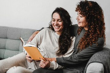 LGBT Hispanic lesbian couple reading a book wearing pajamas in sofa at home - Gay love relationships concept - Focus on left woman face