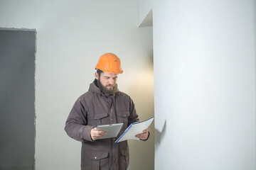 a young man with a beard is standing in an orange construction helmet with a folder of papers and a tablet in his hands.