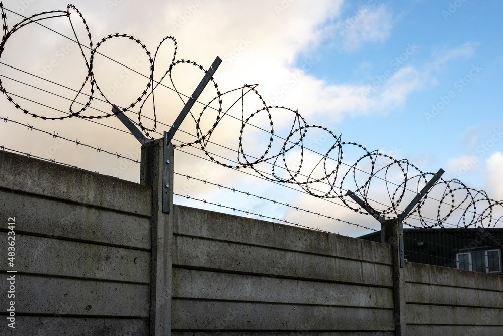 silhouette of security razor wire and barbed wire on top of a panel wall blue sky and clouds 