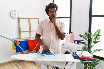 Poster - African man with curly hair ironing clothes at home with hand on chin thinking about question, pensive expression. smiling and thoughtful face. doubt concept.