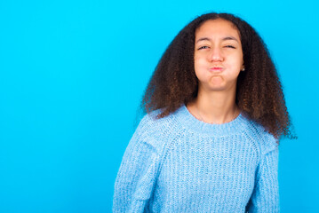 African teenager girl wearing blue sweater over blue background puffing cheeks with funny face. Mouth inflated with air, crazy expression.