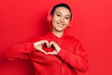 Beautiful hispanic woman with short hair listening to music using headphones smiling in love doing heart symbol shape with hands. romantic concept.