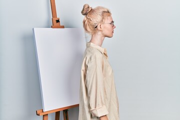 Canvas Print - Beautiful caucasian woman with blond hair standing by painter easel stand looking to side, relax profile pose with natural face with confident smile.