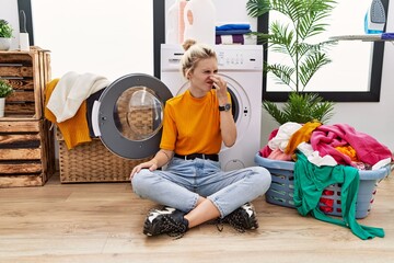 Sticker - Young blonde woman doing laundry sitting by washing machine smelling something stinky and disgusting, intolerable smell, holding breath with fingers on nose. bad smell