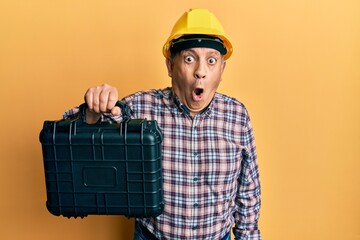 Poster - Handsome senior man with grey hair wearing safety helmet holding toolbox scared and amazed with open mouth for surprise, disbelief face