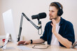 © Girts - Man recording podcast in white studio. hand holding mobile phone in audio home studio. Male with headphones doing online meeting. Remote radio host broadcasting from home with professional microphone.
