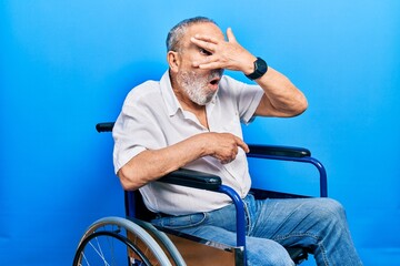 Canvas Print - Handsome senior man with beard sitting on wheelchair peeking in shock covering face and eyes with hand, looking through fingers with embarrassed expression.