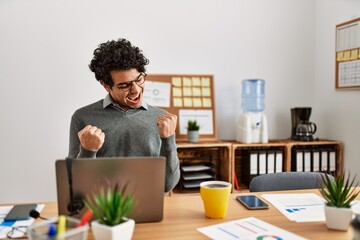 Canvas Print - Young hispanic man wearing business style sitting on desk at office very happy and excited doing winner gesture with arms raised, smiling and screaming for success. celebration concept.