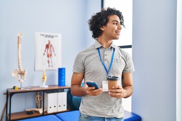 Wall Mural - Young hispanic man physiotherapist using smartphone and drinking coffee at rehab clinic