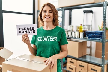 Canvas Print - Middle age woman wearing volunteer t shirt holding please donate banner looking positive and happy standing and smiling with a confident smile showing teeth