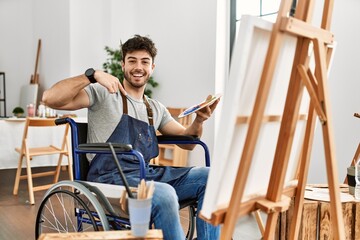 Sticker - Young hispanic man sitting on wheelchair painting at art studio looking confident with smile on face, pointing oneself with fingers proud and happy.