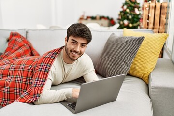 Poster - Young hispanic man smiling happy lying on the sofa using laptop at home.