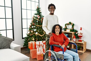 Poster - Young interracial couple with woman sitting on wheelchair by christmas tree sticking tongue out happy with funny expression. emotion concept.