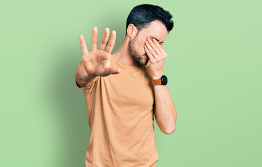 Poster - Hispanic man with beard wearing casual t shirt covering eyes with hands and doing stop gesture with sad and fear expression. embarrassed and negative concept.