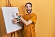 Caucasian man with beard standing drawing with palette by painter easel stand sticking tongue out happy with funny expression.