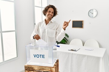 Poster - Young hispanic man voting putting envelop in ballot box with a big smile on face, pointing with hand finger to the side looking at the camera.