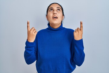 Poster - Young hispanic woman standing over isolated background amazed and surprised looking up and pointing with fingers and raised arms.