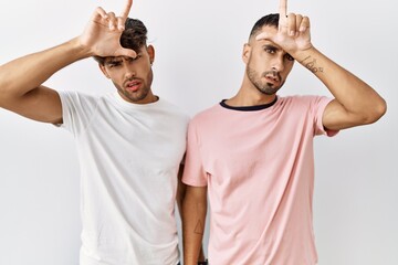 Poster - Young gay couple standing over isolated background making fun of people with fingers on forehead doing loser gesture mocking and insulting.