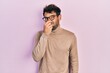 Handsome man with beard wearing turtleneck sweater and glasses smelling something stinky and disgusting, intolerable smell, holding breath with fingers on nose. bad smell