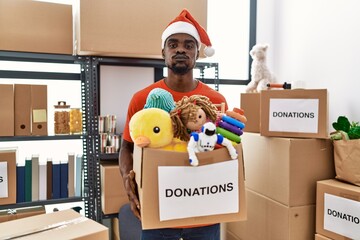 Wall Mural - Young african man wearing volunteer t shirt and christmas hat holding donations puffing cheeks with funny face. mouth inflated with air, catching air.