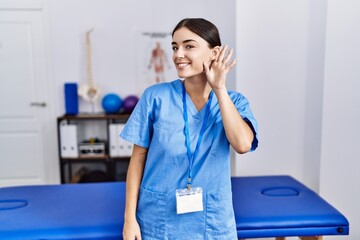 Wall Mural - Young hispanic woman wearing physiotherapist uniform standing at clinic smiling with hand over ear listening an hearing to rumor or gossip. deafness concept.