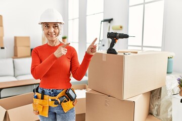 Poster - Middle age grey-haired woman wearing hardhat standing at new home smiling and looking at the camera pointing with two hands and fingers to the side.