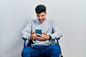 Canvas Print - Young hispanic man sitting on wheelchair using smartphone angry and mad screaming frustrated and furious, shouting with anger. rage and aggressive concept.