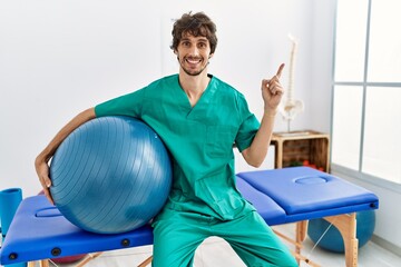 Sticker - Young hispanic physiotherapist man holding pilates ball at pain recovery clinic smiling happy pointing with hand and finger to the side