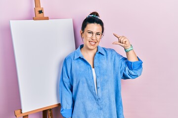 Canvas Print - Young hispanic woman standing by painter easel stand smiling and confident gesturing with hand doing small size sign with fingers looking and the camera. measure concept.