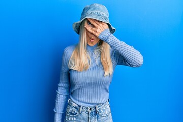 Wall Mural - Young blonde woman wearing casual denim hat peeking in shock covering face and eyes with hand, looking through fingers with embarrassed expression.