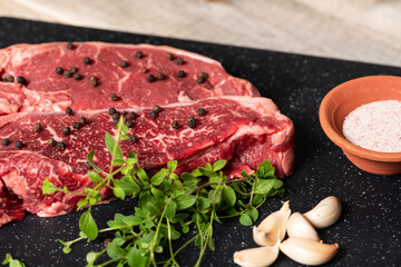 Two raw marbled beef steaks, ready for cooking, with fresh oregano, garlic, and peppercorns.