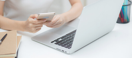 Wall Mural - Closeup hand of young woman working laptop computer and reading smartphone on internet online on desk at home, freelance girl using phone with social media, business and communication concept.