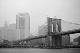 Fototapeta  - Black and white photo of skyscrapers of Manhattan and Brooklyn bridge on foggy and cloudy day. Famous bridge. Postcard view of New York, USA.
