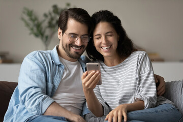 Wall Mural - Millennial couple sit on sofa looking at cellphone screen watching vlog laughing over funny internet content, spend carefree leisure at home enjoy new mobile app. Generation, modern tech usage concept