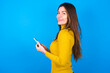 Rear view photo portrait of young woman wearing yellow turtleneck sweater against blue background using smartphone smiling