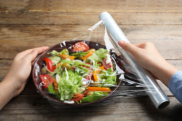 Canvas Print - Woman putting plastic food wrap over bowl of fresh salad at wooden table, closeup