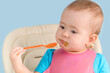 © Larisa - A baby 12-17 months old in a pink bib eats mashed broccoli with a spoon while sitting on a feeding chair. Close-up