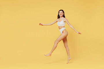 Wall Mural - Full body side view young smiling fun brunette woman 20s wearing white underwear with perfect fit figure walking going srolling look camera isolated on plain yellow color background studio portrait.