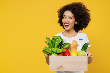 Young happy smiling amazed woman of African American ethnicity wears white t-shirt hold box with vegetarian vegan food organic vegetables look aside on workspace isolated on plain yellow background